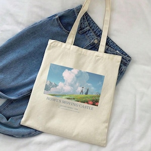 May include: Cream-colored tote bag featuring a colorful landscape scene with two figures and the text "Howl's Moving Castle". The bag has long straps and is placed on top of blue denim jeans.