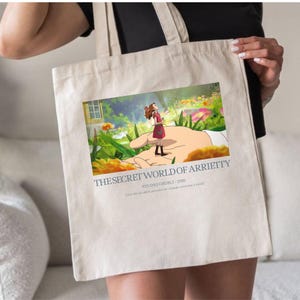 K&ouml;nnte beinhalten: Beige Tragetasche mit einer farbenfrohen Illustration aus "Arrietty &ndash; Die kleine Welt der Borger". Das Bild zeigt ein M&auml;dchen in einem roten Kleid, das auf einer riesigen Hand in einem Garten steht. Text enth&auml;lt Filmtitel und "Studio Ghibli 2010."