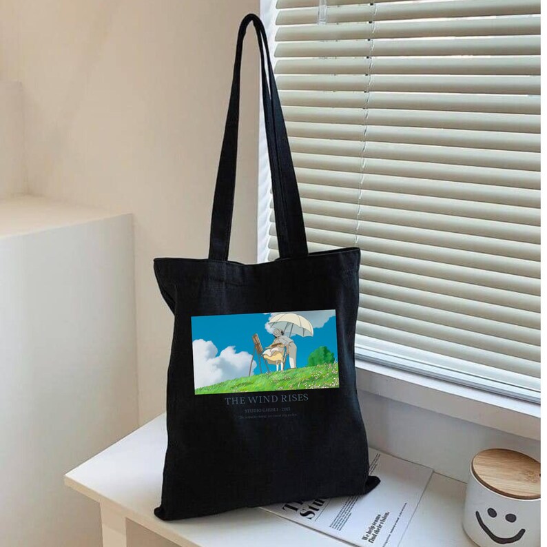 May include: Black tote bag with long handles. The bag features a colorful graphic of a cartoon scene with the text "THE WIND RISES". The bag is made of a sturdy material and is suitable for carrying various items.
