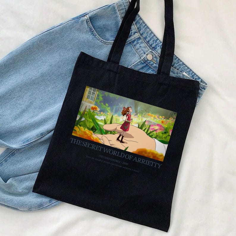 K&ouml;nnte beinhalten: Schwarze Tragetasche mit einer farbenfrohen Illustration aus "Arrietty &ndash; Die wundersame Welt der Borger". Das Bild zeigt ein M&auml;dchen, das auf einer riesigen Hand in einem Garten steht. Der Text "THE SECRET WORLD OF ARRIETTY" ist darunter gedruckt. Die Tasche hat schwarze Griffe.