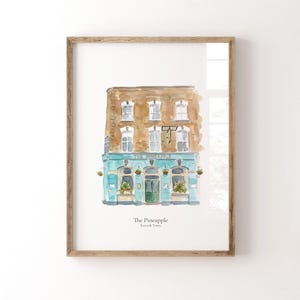May include: A framed watercolor art print depicting a building facade with a brown brick upper level and a teal ground floor. The text "The Pineapple" and "Kentish Town" is printed below the illustration of the pub with arched windows.