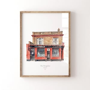 May include: A framed watercolor print depicting The Boogaloo pub in Highgate, N6. The building features a brick upper story with two windows and a vibrant red ground floor with dark trim, large windows, and a central door. The text reads The Boogaloo, Highgate, N6.