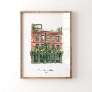 Op de afbeelding: Ingelijste aquarelprint van The Devonshire pub in Soho. Het rode bakstenen gebouw heeft groene afwerking en ramen en is bedekt met groen. De tekst "The Devonshire Soho" staat onderaan de afbeelding.