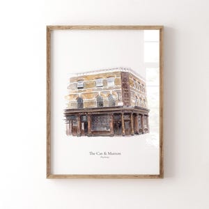 May include: A framed watercolour print depicting The Cat & Mutton public house in Hackney. The building features a tan brick facade, dark brown ground floor storefront, and arched windows. The text 'The Cat & Mutton' and 'Hackney' appear below.