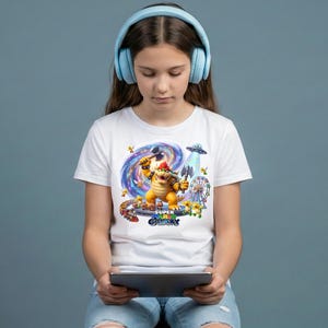May include: A person wearing light blue headphones and a white t-shirt with a colourful graphic of a video game character. The graphic includes the text "SUPER GALAXY". The person is holding a tablet.