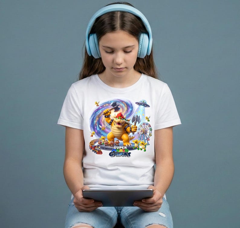 May include: A person wearing light blue headphones and a white t-shirt with a colourful graphic of a video game character. The graphic includes the text "SUPER GALAXY". The person is holding a tablet.