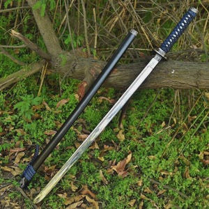 Op de afbeelding: Een Japanse katana-zwaard met een zwarte schede en een blauw-witte handgreep. Het zilveren lemmet ligt op een bed van groen gebladerte en gevallen bladeren.