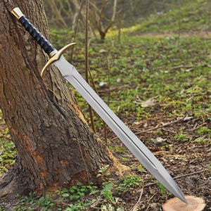 Pode incluir: Uma espada longa e prateada com uma lâmina de dois gumes, encostada a uma árvore. O punho apresenta uma pega preta, detalhes dourados e um pomo dourado. A lâmina da espada tem cerca de 91 cm de comprimento.