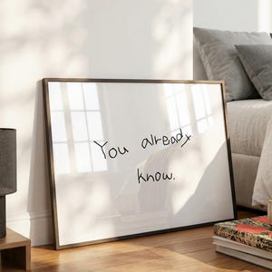 以下が含まれることがあります： 「You already know」と手書きされたテキストが入った額入りの白いボード。フレームは濃い茶色です。ボードは寝室の白い壁に立てかけられています。本とマグカップの山が前景にあります。