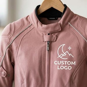 Close-up shot of the upper torso of a dusty pink ladies' leather motorcycle jacket. The image highlights the premium cowhide texture, white piping details, shoulder padding, and a sharp white geometric moon-and-mountain logo with the text "CUSTOM LOGO" printed on the chest.