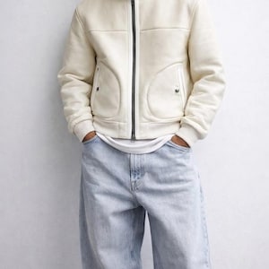 May include: Cream-colored jacket with a black zipper and two front pockets. The jacket has a high collar and ribbed cuffs. The model is wearing light blue jeans.