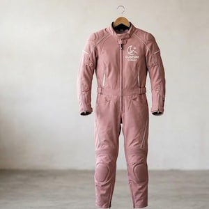 Front full-length view of a minimalist pink women's cowhide leather riding suit hanging on a wooden hanger inside a motorcycle gear shop. The sleek one-piece suit features integrated knee pads, a central zipper, and a white minimalist custom logo printed on the left chest.
