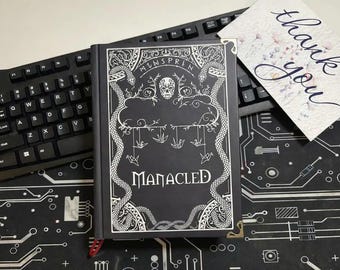 May include: A black hardcover book titled Manacled with white line art featuring a skull, two snakes, and hanging origami cranes. The book has gold-toned metal corner protectors and a red ribbon bookmark. A thank you card is placed nearby.