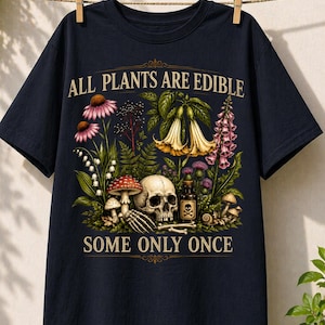 May include: Navy blue t-shirt with a botanical and skull design. The graphic features various flowers, mushrooms, a skull, and the text "ALL PLANTS ARE EDIBLE SOME ONLY ONCE". The shirt is hanging on a clothesline.
