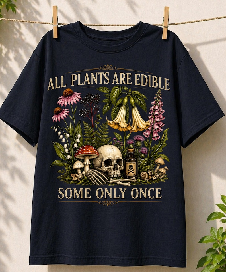 May include: Navy blue t-shirt with a botanical and skull design. The graphic features various flowers, mushrooms, a skull, and the text "ALL PLANTS ARE EDIBLE SOME ONLY ONCE". The shirt is hanging on a clothesline.
