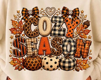 Könnte beinhalten: Ein cremefarbenes Sweatshirt mit Rundhalsausschnitt und dem Schriftzug COZY SEASON in großen, gemusterten Blockbuchstaben. Das Design zeigt Leopardenmuster, Karos und Punkte, verziert mit Herbstblättern, Eicheln und zwei karierten Schleifen.