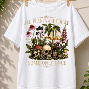 May include: White t-shirt with a botanical illustration featuring flowers, mushrooms, a skull, and a poison bottle. The text "ALL PLANTS ARE EDIBLE" and "SOME ONLY ONCE" are printed in gold.
