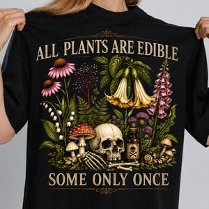 May include: Black t-shirt featuring a botanical design with the text "ALL PLANTS ARE EDIBLE SOME ONLY ONCE." The illustration includes flowers, mushrooms, a skull, and a poison bottle.