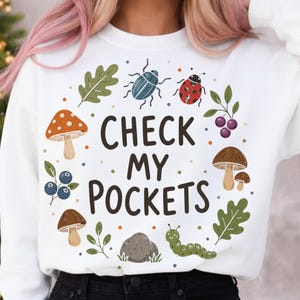 May include: White sweatshirt with a playful design. It features illustrations of mushrooms, leaves, a ladybug, and a caterpillar, along with the text "CHECK MY POCKETS". The design is encircled by colorful dots.