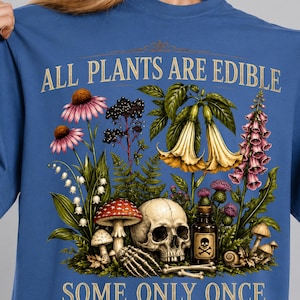 May include: Blue t-shirt with a botanical illustration featuring flowers, mushrooms, a skull, and the text "ALL PLANTS ARE EDIBLE, SOME ONLY ONCE". The design includes a poison bottle and a skeleton hand.