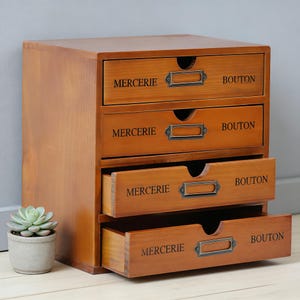 May include: A wooden storage cabinet with four drawers, each labeled with the words "MERCERIE" and "BOUTON" in black text. The cabinet has a warm, brown finish and metal drawer pulls. One drawer is partially open, revealing its interior.