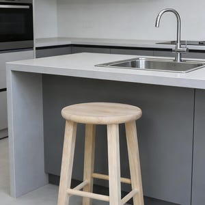 May include: A light-colored wooden stool with a round seat and four straight legs. The stool is positioned in front of a kitchen island with a stainless steel sink and faucet. The kitchen features gray cabinets and countertops.