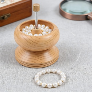 May include: A wooden bead spinner with a metal needle, filled with white and gold beads. A partially completed pearl bracelet is in the foreground. A magnifying glass and jewelry box are in the background.