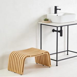 May include: A light-colored wooden stool with a curved design, constructed from vertical slats. The stool is near a white sink and a black metal stand. The bathroom fixture has a black faucet and a small succulent plant.