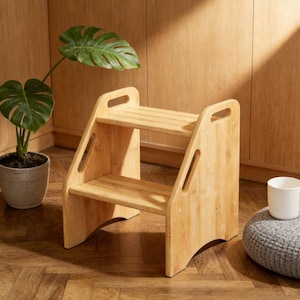 May include: A two-step wooden step stool made of light-colored bamboo. The stool has a rectangular shape with two steps and handles cut into the sides. The stool is approximately 18 inches tall and 16 inches wide.