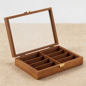 May include: A wooden box with a clear glass lid, open to reveal eight compartments. The box is made of a warm-toned wood and has a brass-colored clasp. The interior is lined with a brown felt material. The box is designed for organizing small items.