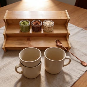 May include: A wooden tiered shelf holds three small jars filled with green powder, brown granules, and oats. Two cream-colored mugs are stacked, and another sits beside a long copper spoon. The items are arranged on a light-colored linen cloth.
