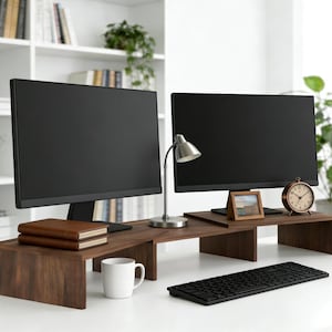 May include: A home office setup featuring two black computer monitors on a dark brown wooden desk. A small silver desk lamp, a vintage-style clock, and a keyboard are also visible. A white mug and two books are on the desk.