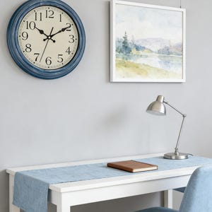 May include: A blue-framed wall clock with a cream-colored face and black numerals hangs on a gray wall. A watercolor landscape painting in a white frame is next to the clock. A silver desk lamp and a brown book sit on a white desk with a blue runner.