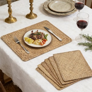 May include: A dining table setting featuring woven placemats and matching coasters. A plate of food with a steak, pasta, and vegetables sits on a placemat. Wine glasses and antique candle holders complete the scene.