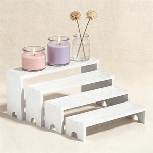 May include: A white wooden tiered display stand with four steps. Two lit candles in glass jars, one pink and one purple, sit on the top step. A small glass jar with dried flowers is also on the top step. The stand is made of white-painted wood.