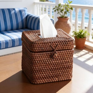 May include: A square, woven rattan tissue box in a warm brown hue. The box has a lid and a small opening for tissue dispensing. White tissues are visible. The box is on a wooden surface, with a coastal-themed background.