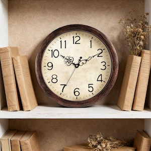 May include: A round wall clock with a brown frame and a cream-colored face. The clock features black numerals and hands, and is surrounded by several books and dried flowers, creating a vintage aesthetic. The clock is displayed on a white shelf.