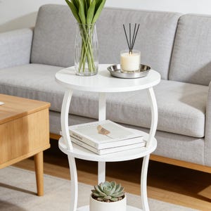 May include: A white, two-tiered side table with a round top and bottom shelf. The top shelf holds a vase of white tulips, a candle, and a small metal tray. The bottom shelf displays a potted succulent and a stack of books.