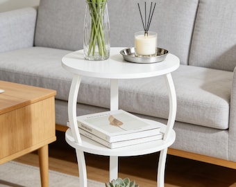 May include: A white, two-tiered side table with a round top and bottom shelf. The top shelf holds a vase of white tulips, a candle, and a small metal tray. The bottom shelf displays a potted succulent and a stack of books.