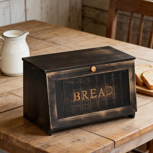 May include: A black wooden bread box with the word "BREAD" in gold lettering. The box has a distressed finish and a wooden knob. It sits on a wooden table, with a white pitcher and slices of bread in the background.