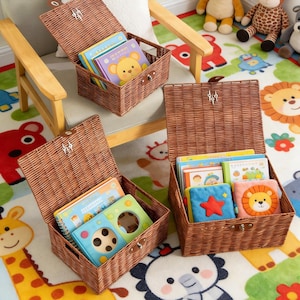May include: Three open wicker storage baskets filled with children's books. The baskets are brown and rectangular, with hinged lids. The books have colorful covers and are stacked inside the baskets. The baskets are on a colorful rug with animal designs.