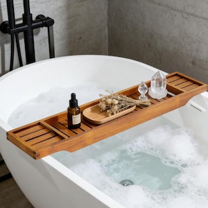 May include: A wooden bathtub tray with a dark finish, holding a small wooden tray with dried flowers, a bottle with a dropper, and two clear crystal decorations. The tray rests on the edge of a white bathtub filled with soapy water.