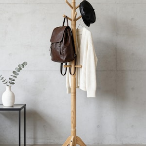 May include: A wooden coat rack with a brown leather backpack, a white sweater, and a black beret. The rack has multiple pegs for hanging items and a tripod base. The background is a concrete wall and a patterned rug.