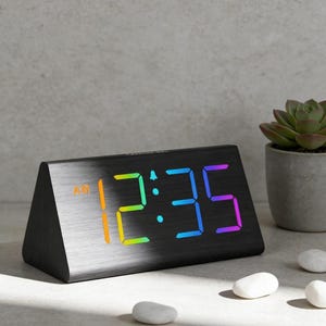 May include: Black digital alarm clock displaying 12:35 AM with a rainbow gradient. The clock has a brushed metal finish and a triangular shape. A small succulent plant in a gray pot and white stones are in the background.