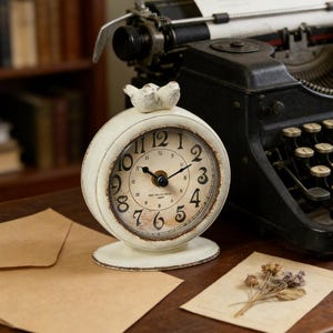 May include: Cream-colored, vintage-style clock with a distressed finish. The clock features black numerals, a black clock hand, and a decorative bird on top. The clock is set on a wooden surface, next to a vintage typewriter and stationary.