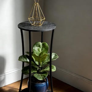 May include: A black and gray plant stand with a round top and a lower shelf. A blue ceramic pot with a green plant sits on the lower shelf. A gold geometric vase with branches sits on top.