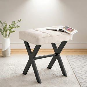 May include: A beige upholstered ottoman with a tufted top and black metal X-shaped legs. A magazine rests on the ottoman. The ottoman is on a light-colored rug.