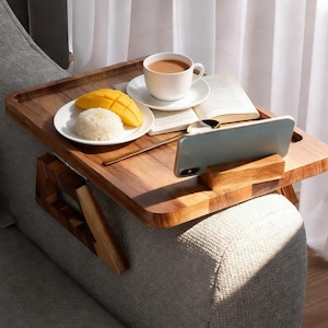 May include: A wooden sofa arm tray with a built-in phone holder. The tray holds a white plate with mango and rice, a cup of coffee, and an open book. The tray is attached to a gray sofa.