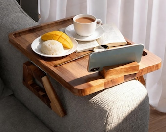 May include: A wooden sofa arm tray with a built-in phone holder. The tray holds a white plate with mango and rice, a cup of coffee, and an open book. The tray is attached to a gray sofa.