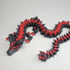 Könnte beinhalten: Ein 3D-gedrucktes, bewegliches Drachenspielzeug in Schwarz und Rot. Der Drache hat ein detailliertes, stacheliges Design mit einem segmentierten Körper, der eine flexible Pose ermöglicht. Kopf und Schwanz sind klar definiert, und das Farbschema ist auffällig.